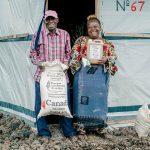 A man and a woman stand in front of a tent shelter holding sacks labeled as food aid from Canada.