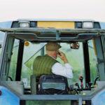 Senior male farmer driving tractor, using mobile phone, rear view