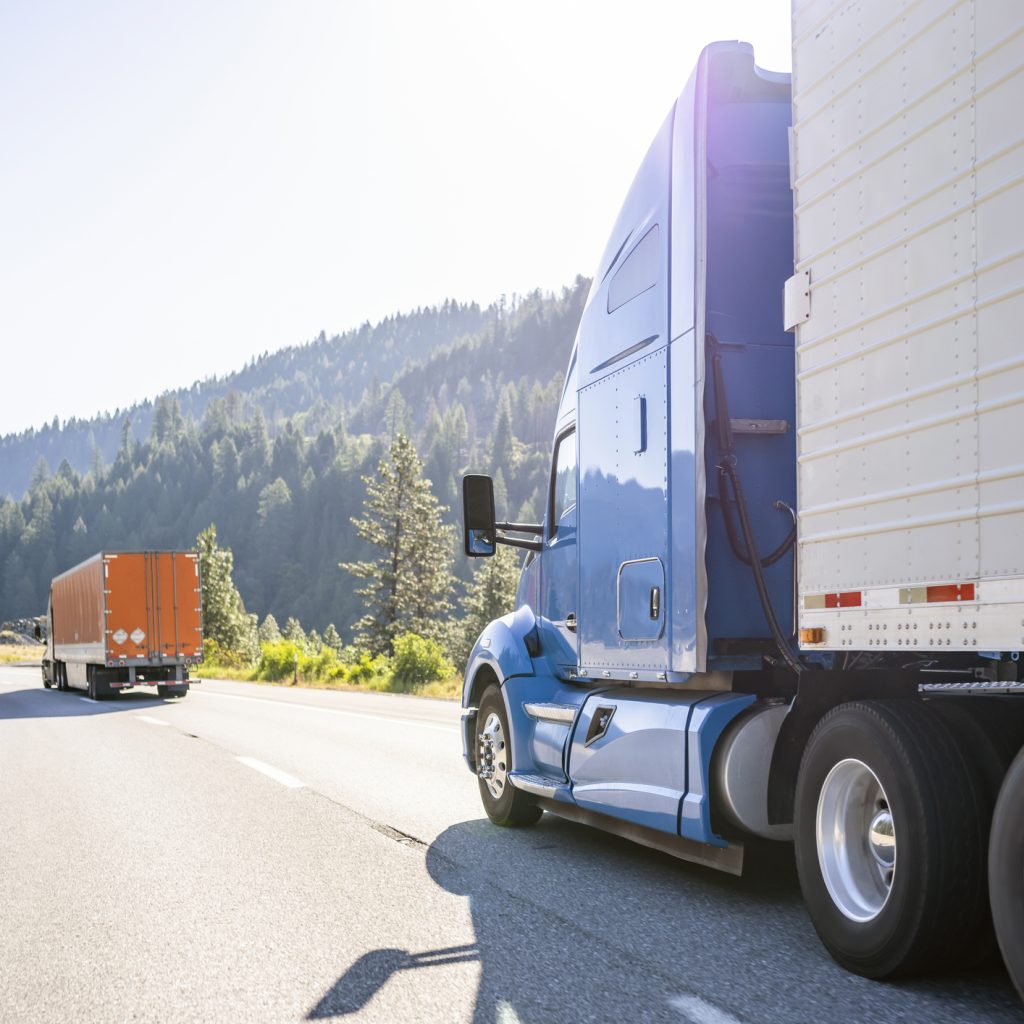 A blue semi truck in the foreground drives down a highway with a forest in the background.