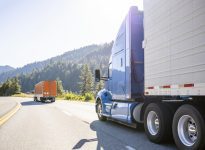 A blue semi truck in the foreground drives down a highway with a forest in the background.