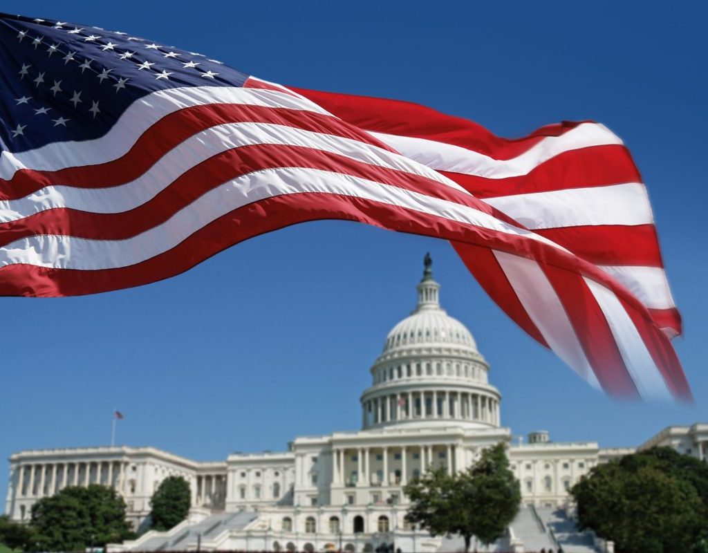 The U.S. flag flies in the foreground with the Capitol building in the background, framed by the flag.