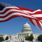The U.S. flag flies in the foreground with the Capitol building in the background, framed by the flag.