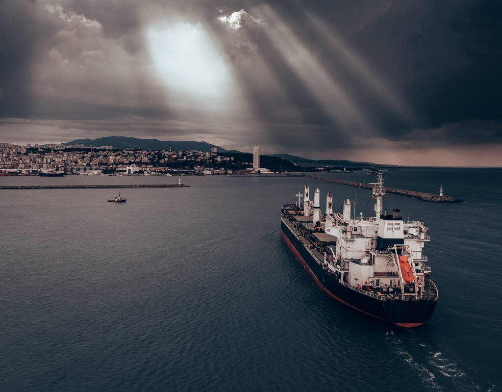 A cargo ship approaches port under a cloudy sky.