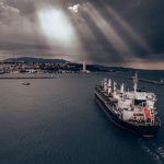 A cargo ship approaches port under a cloudy sky.