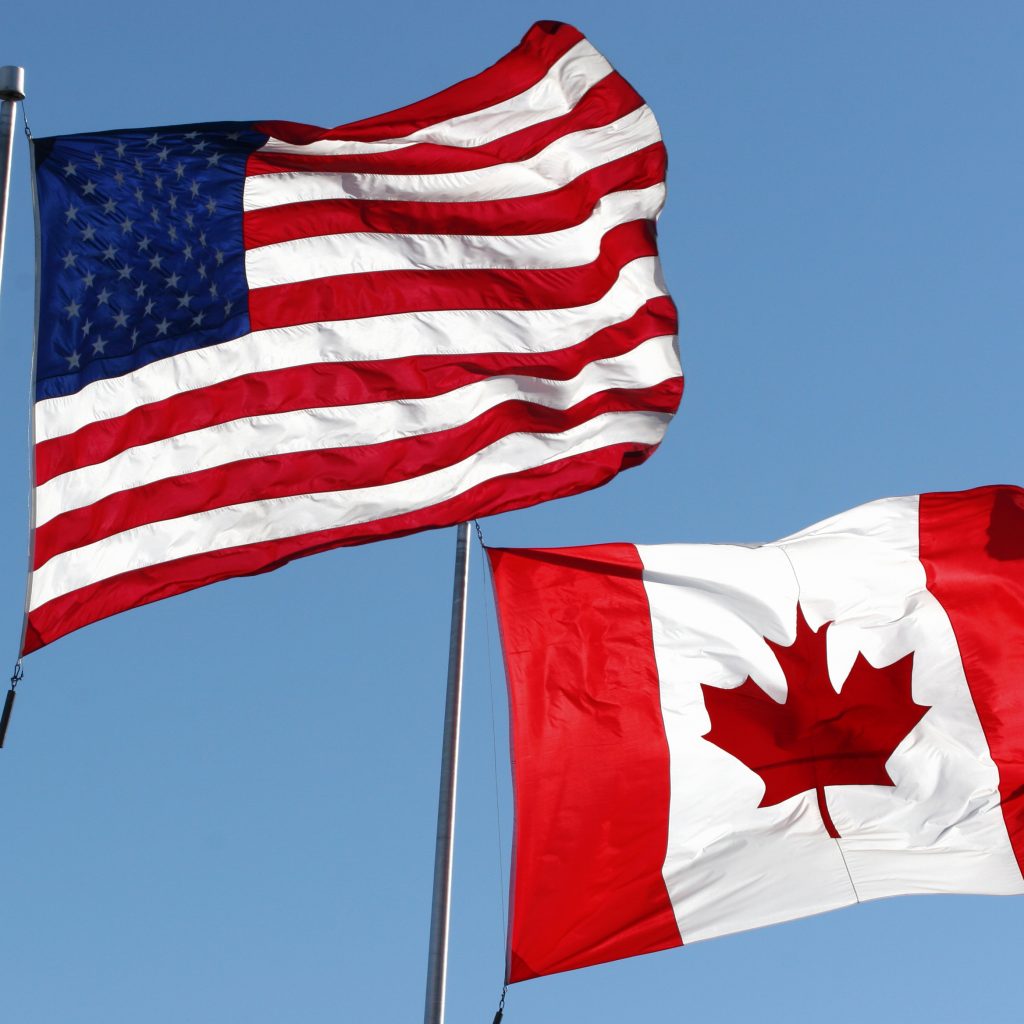 An American and Canadian flag fly side by side in a blue sky.