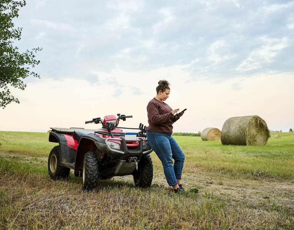 Female farmer using mobile phone while standing by an ATV on the farm field with hay bales