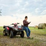 Female farmer using mobile phone while standing by an ATV on the farm field with hay bales