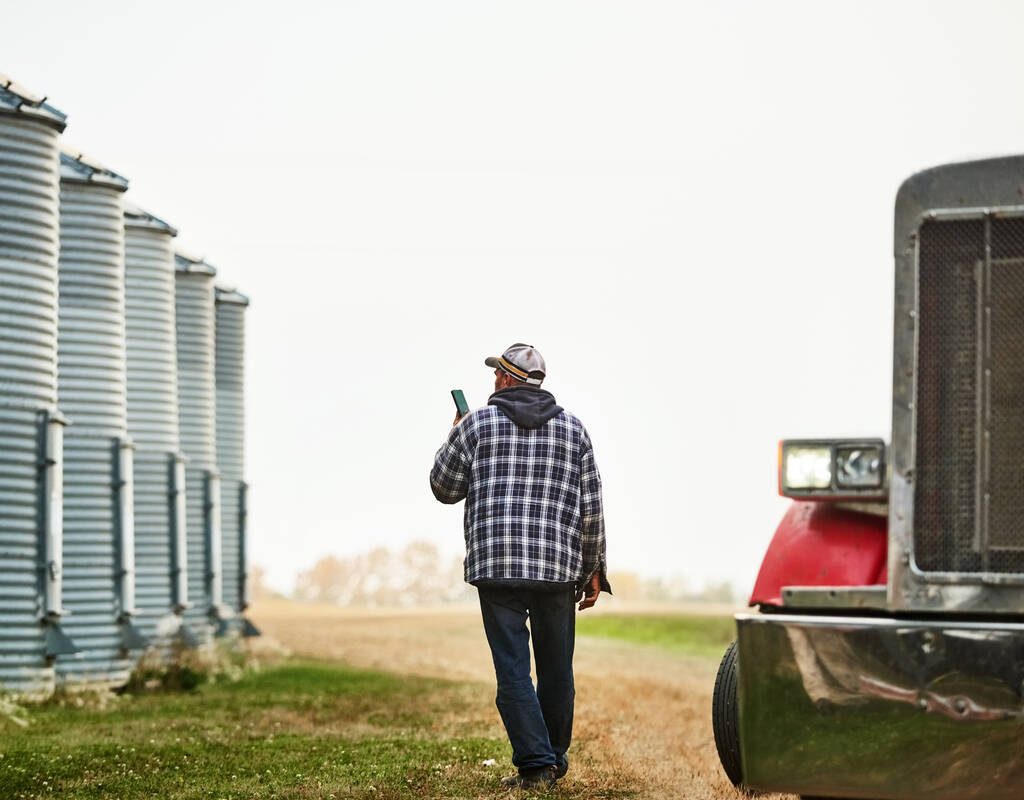 Rear view of a male farm worker with phone walking outside along the large grain storage silos on farm. Photo: Getty Images Plus