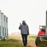 Rear view of a male farm worker with phone walking outside along the large grain storage silos on farm. Photo: Getty Images Plus