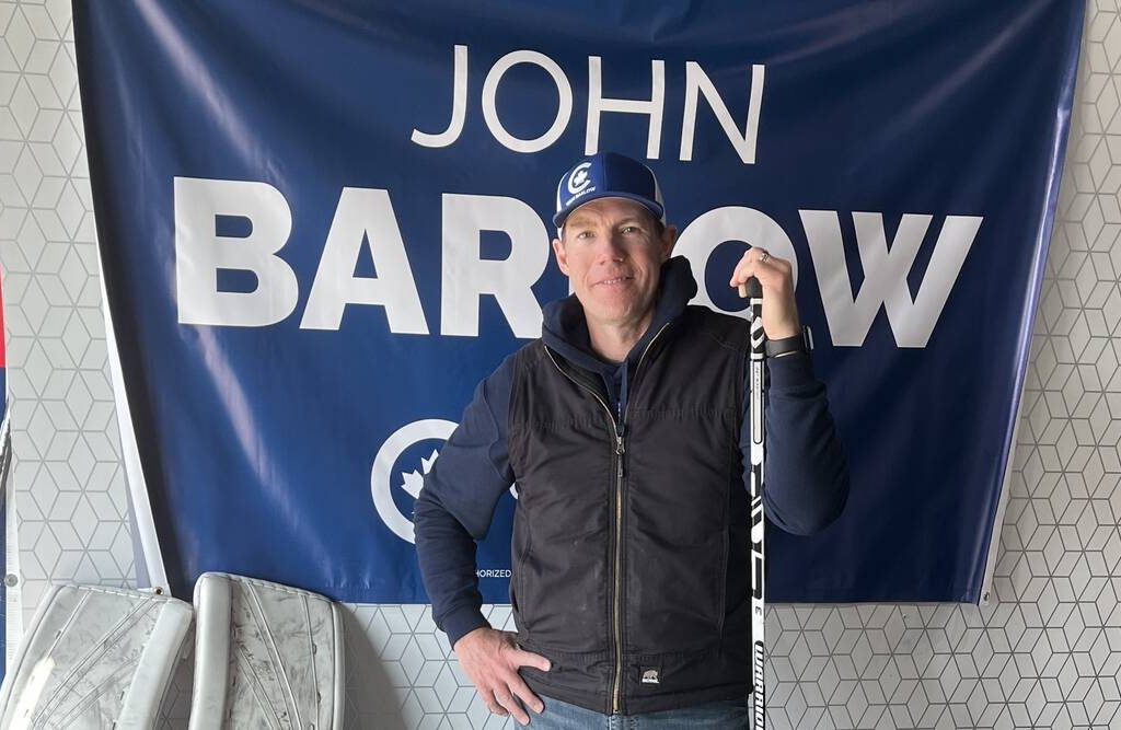 John Barlow standing in front of a campaign flag and holding a hockey stick. Photo; Greg Price
