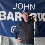John Barlow standing in front of a campaign flag and holding a hockey stick. Photo; Greg Price