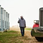Rear view of a male farm worker with phone walking outside along the large grain storage silos on farm. Photo: Getty Images Plus