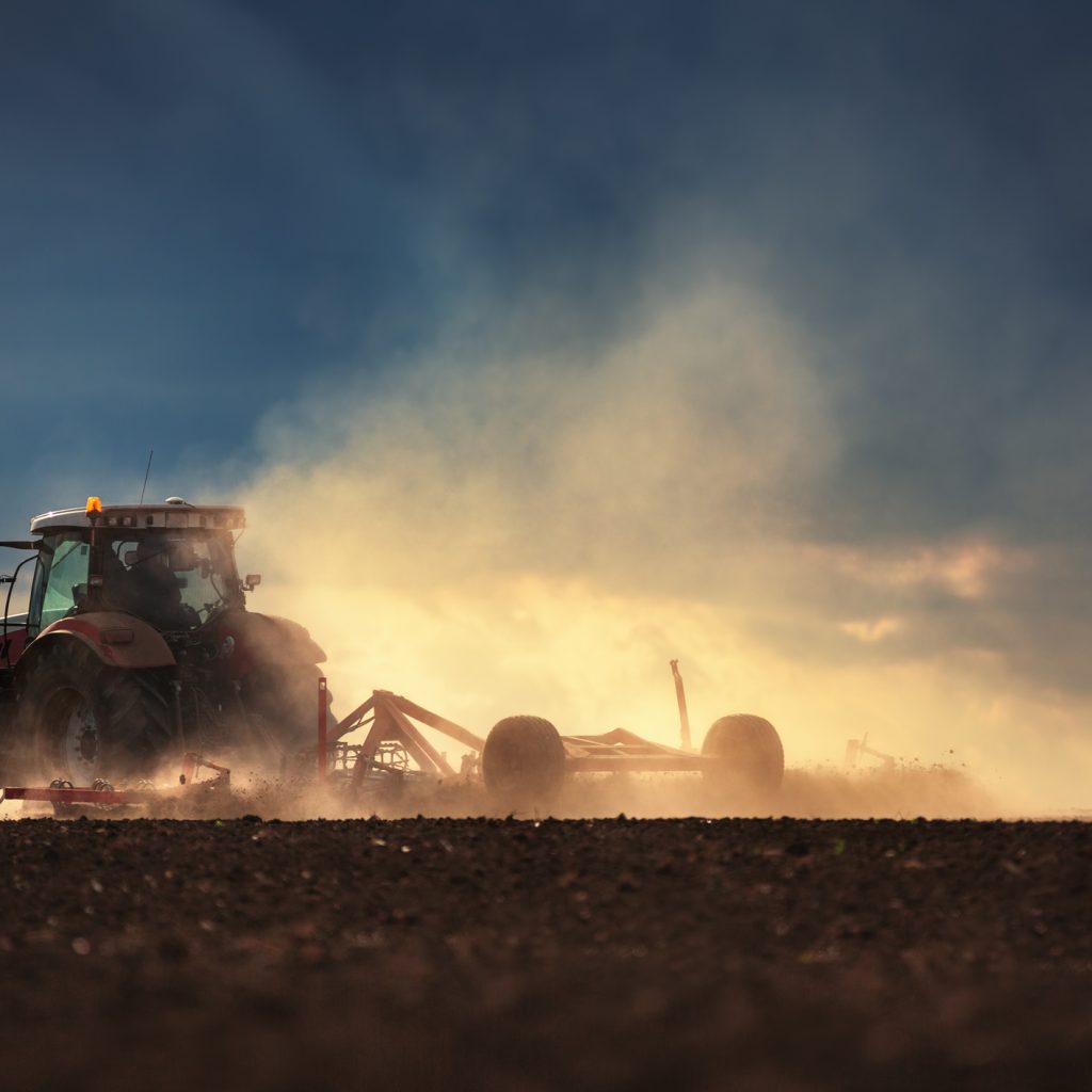 A tractor pulls a cultivator or harrow through a dust cloud.