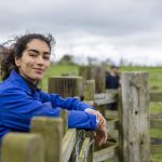 A portrait of a young female farmer wearing overalls, standing and leaning against a fence at the farm she works at.