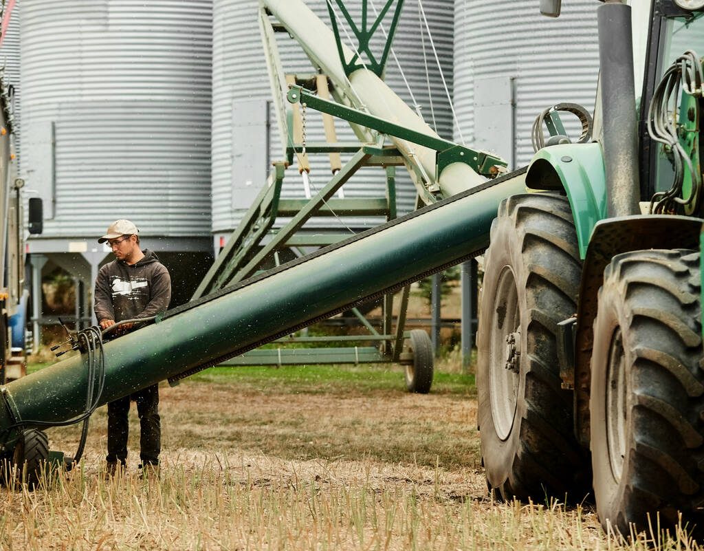 Young man working on farm operating grain auger alongside grain bins. Photo: AJ_Watt/Getty Images Plus