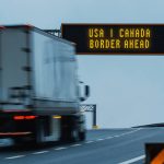 Semi truck approaching a border ahead highway sign.