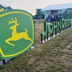 A John Deere logo stands in an outdoor, grassy area at a farm show.