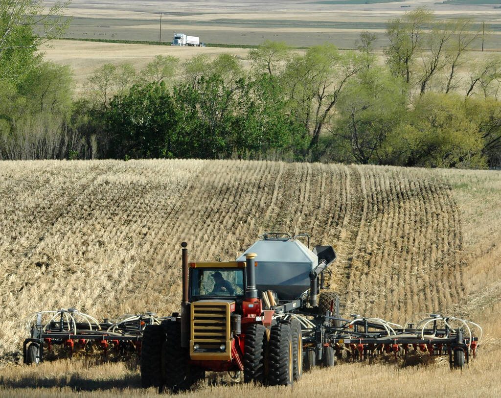 A large red tractor pulls an air seeder rig over a hill in a Prairie field. It is seeding directly into stubble.