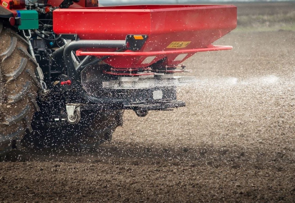 A red fertilizer mounted on the back of a tractor broadcasts white granules of fertilizer on a cultivated field.