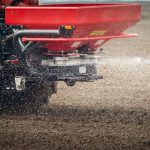 A red fertilizer mounted on the back of a tractor broadcasts white granules of fertilizer on a cultivated field.
