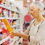 A woman stands in a grocery store reading the label on a cereal box.